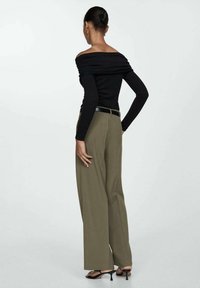 Vrouw staat met de rug naar de camera, draagt een zwarte off-shoulder langemouwen top, olijfgroene brede broek, zwarte riem en zwarte sleehakken.