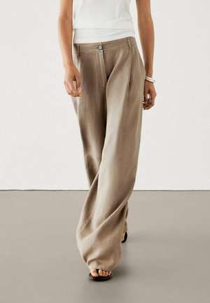 Femme portant un pantalon en lin beige à taille haute et jambes larges avec bouton et fermeture éclair, associé à un haut blanc sans manches et des sandales noires.