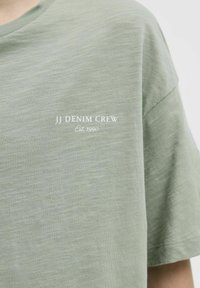 Gaiši zaļa kokvilnas T-kreklu ar kārtīgu apkakli, uz kreisā krūts ir balts uzraksts "JJ DENIM CREW" un "Est 1990". Audums ar struktūru.