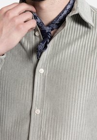 Camisa de color verde claro, con canalé y un collar clásico, que presenta un cierre de botones y que se combina con un pañuelo de paisley en tonos azul marino alrededor del cuello.