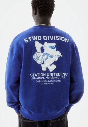 Osoba nosící modrou mikinu s bílým motivem medvěda držícího basketbalový míč a vlajku, s textem "STWD DIVISION" a "Station United Inc, Maryland, USA"