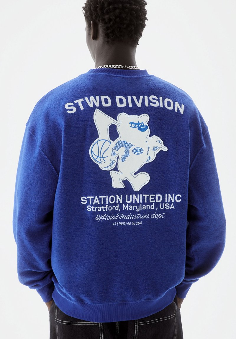Personne portant un sweat-shirt bleu avec un motif blanc représentant un ours tenant un ballon de basket et un drapeau, avec le texte "STWD DIVISION" et "Station United Inc, Maryland, USA"