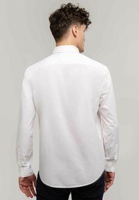 Biała koszula zapinana na guziki z klasycznym kołnierzykiem, długimi rękawami i gładką fakturą. Posiada zaokrąglony dół oraz mankiety zapinane na guziki.