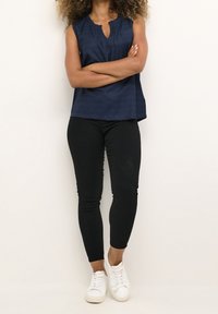 Marinblå ärmlös topp med v-hals och texturerat tyg, i kombination med svarta leggings och vita sneakers; armarna i kors. Neutral bakgrund.