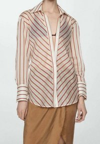 Camicia a maniche lunghe in tessuto trasparente con righe diagonali rosse e bianche, dotata di colletto classico e chiusura a bottoni, abbinata a una gonna marrone.