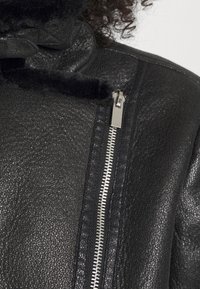 Giubbotto in pelle nera con superficie texturizzata, dotato di chiusura con zip metallica e colletto foderato in pelliccia. Gli accenti includono un dettaglio decorativo con zip.