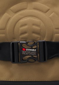Correas frontales de patineta plegables en tejido marrón con un patrón texturizado. Presenta una hebilla ajustable negra y una etiqueta con el logo para identificación.