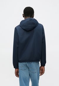 Ελαφρύ μπουφάν σε ναυτικό μπλε χρώμα με κουκούλα, ελαστικές μανσέτες και κομμένη μέση. Λεία υφή, μίνιμαλ σχεδιασμός, συνδυασμένο με τζιν σε ανοιχτό μπλε.