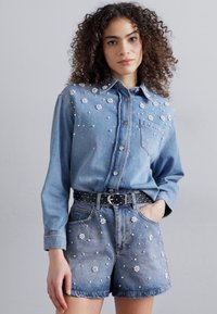 Camicia di denim con applicazioni di fiori decorativi e bottoni, abbinata a pantaloni corti coordinati. Una cintura nera con borchie mette in risalto la vita.