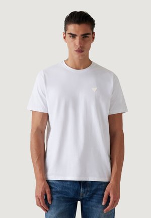 Uomo con capelli pettinati all'indietro che indossa una maglietta bianca semplice a girocollo e jeans blu, in piedi davanti a uno sfondo neutro chiaro.