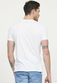 T-shirt blanc à manches courtes, avec un col rond. Matière en coton doux avec des coutures minimales. La vue de dos montre un petit logo près du col.