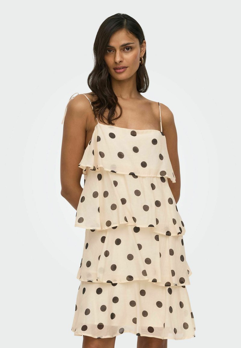 Femme aux cheveux foncés portant une robe beige à volants avec des pois noirs et des bretelles fines, debout devant un fond clair uni.