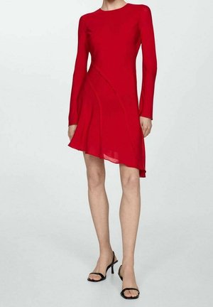 Kobieta w czerwonej, asymetrycznej sukience z długim rękawem oraz czarnych, odkrytych sandałach na wysokim obcasie, stojąca na jednolitym tle.