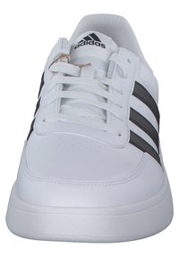 Vit sportskor med en slät syntetisk läderöverdel, som har tre svarta ränder och en vadderad plös. Snörena går igenom åtta snörhål.