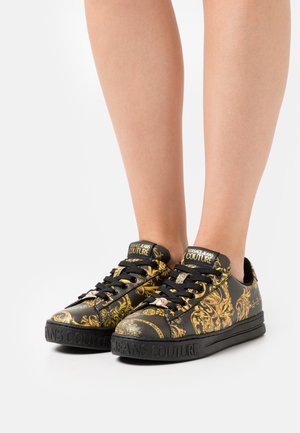 Svarta sneakers med ett gyllene barockmönster, texturerat material, rund tå, svarta skosnören och branding på tungan och sulan.