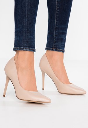 Beige leather high heels with a pointed toe and a slender, stiletto heel, paired with dark blue denim jeans. Smooth texture and minimal design.