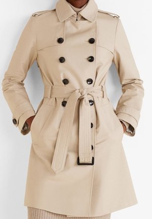 Beige dubbelrijige trenchcoat met zwarte knopen, bijpassende riem gestrikt in de taille, knoopmanchetten en handen in de zijzakken.
