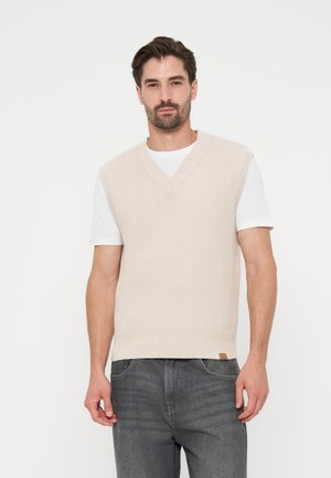 Mann mit dunklen Haaren und Bart, der einen beigen gestrickten ärmellosen Pullover über einem weißen T-Shirt und grauen Jeans trägt und vor einem weißen Hintergrund posiert.