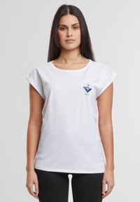 Hvid bomulds t-shirt med rulleærmer, der har et lille blåt geometrisk logo på venstre brystområde. Enkel, afslappet design.