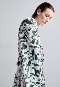 Langärmliges Kleid aus leichtem Stoff mit grün-weißer Blättergestaltung und roten und orangefarbenen Akzenten. Der drapierte Schnitt verstärkt die Fluidität.