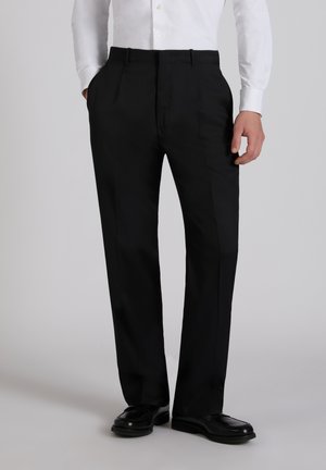 Mężczyzna ubrany w czarne, proste spodnie garniturowe, czarne buty oraz białą, długorękawową koszulę zapinaną na guziki, stojący z jedną ręką w kieszeni.