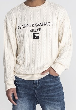 Homme portant un pull en maille torsadée crème avec le texte et le logo "Gianni Kavanagh Atelier", associé à un jean bleu, main dans la poche.