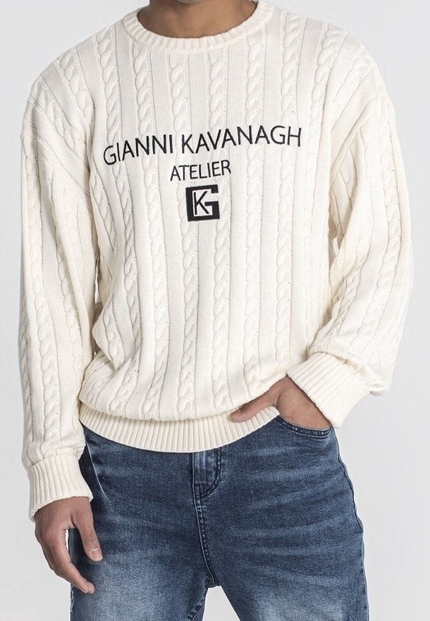 Homme portant un pull en maille torsadée crème avec le texte et le logo "Gianni Kavanagh Atelier", associé à un jean bleu, main dans la poche.