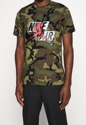 Person trägt ein grünes und braunes Camouflage Nike Air T-Shirt mit schwarzen Hosen und steht vor einem schlichten hellen Hintergrund.