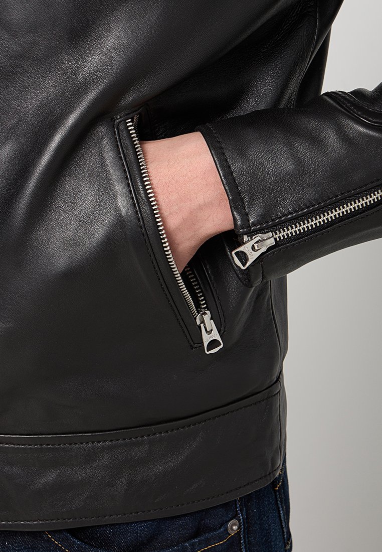 Chaqueta de cuero negra con un bolsillo con cremallera, que presenta una textura suave, un diseño elegante y herrajes metálicos en los puños.
