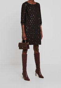 Robe noire avec des motifs métalliques, manches trois quarts, assortie à un sac à main marron et des bottes à talons marron jusqu'aux genoux.