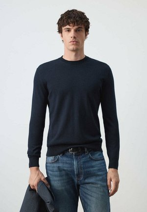 Joven con el pelo corto y rizado, lleva un suéter oscuro de manga larga y vaqueros azules, sosteniendo una chaqueta oscura en su mano izquierda.