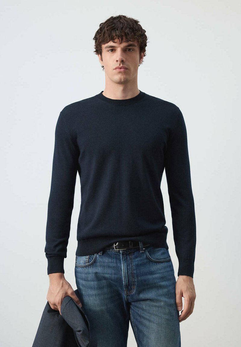 Jeune homme aux cheveux courts et bouclés, portant un pull sombre à manches longues et un jean bleu, tenant une veste sombre dans sa main gauche.