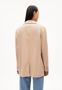 Blazer beige su misura con un design strutturato, caratterizzato da uno spacco posteriore singolo e polsini con bottone, indossato sopra una camicia a scacchi e jeans di denim.
