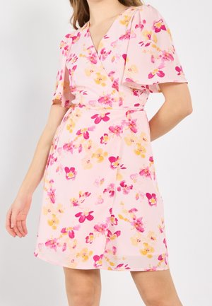 Kvinde iført en pink blomstret slå-om-kjole med korte flagreærmer, prydet med gule og magentarøde blomster.