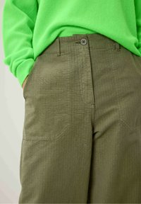 Alyvų žalia kelnės iš tekstūruoto audinio, su priekinėmis kišenėmis ir mygtukų uždarymu. Derinamos su ryškiai žalia, minkštos mezginio megztiniu.