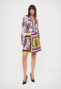 Vrouw in een kleurrijke blouse met vlinder- en bloemenprint, met een bijpassende geplooide rok en zwarte hoge hakken, staande tegen een witte achtergrond.