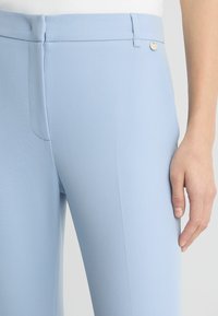 Pantaloni su misura blu chiaro realizzati in tessuto morbido, con una tasca frontale e un piccolo dettaglio con bottone metallico in vita.