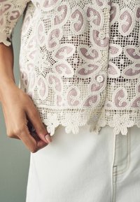 Bluse i blonde med indviklede lilla paisley-mønstre, bølget kant og knapdetaljer. Paret med lyse stoffer nederst.