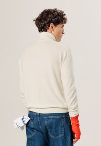 Cremfarbener Strickpullover mit hohem Kragen, kombiniert mit blauen Denim-Jeans. Auffälliges Detail ist der orangefarbene gerippte Ärmelakzent und ein weißes Taschentuch.