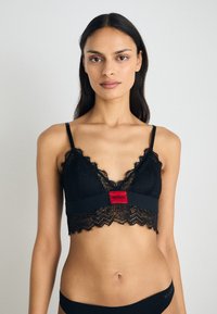 Svart blonde bralette med bølget kant, med et rødt logo bånd. Justerbare stropper og myk tekstur, designet for komfort og stil.