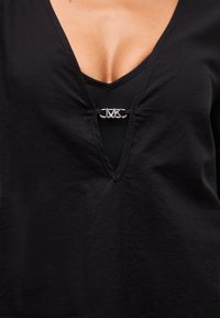 Κοντινό πλάνο μαύρου τοπ με V-λαιμόκοψη και ασημένιο κούμπωμα με το λογότυπο MK που ενώνει το ντεκολτέ στο ύψος του στήθους.