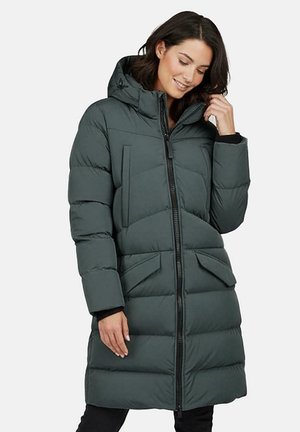 Vrouw draagt een lange donkergroene gewatteerde winterjas met capuchon, rits aan de voorkant en klepzakken, glimlacht en raakt haar haar aan.