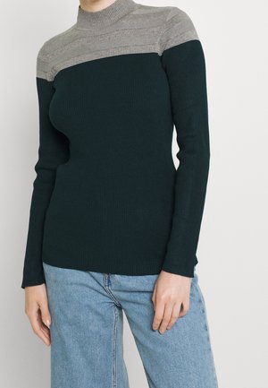 Femme portant un pull côtelé cintré avec un empiècement rayé gris et un corps vert foncé, associé à un jean bleu clair sur un fond uni.
