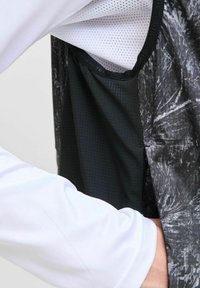 Juodas ir pilkas sportinis liemenė su tekstūruotu raštu, viduje turintis tinklinę pamušalą ir balta ilgomis rankovėmis marškinį po juo.