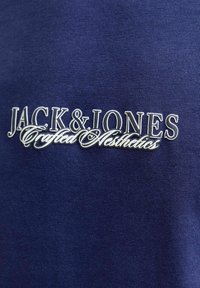 Granatowy materiał z teksturowanym prążkowaniem zawiera wyraźne białe haftowane logo: "JACK & JONES Crafted Aesthetics" z ozdobnymi akcentami.
