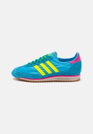 Jasnoniebieski but sportowy Adidas z żółtymi paskami, zielonymi sznurowadłami, różowymi akcentami i gumową podeszwą, pokazany z boku na białym tle.