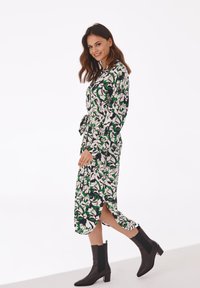Blumenmuster-Kleid mit grünen und weißen Mustern auf schwarzem Hintergrund; lange Ärmel, Taillenband, Schlitzdetails am Saum. Getragen mit schwarzen Knöchelstiefeln.