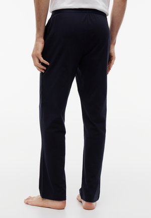 Personne pieds nus portant un pantalon droit bleu marine foncé et une chemise blanche, vue de dos sur un fond uni.