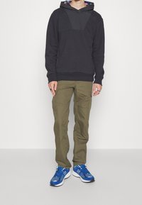 Svart hoodie med en texturerad bröstpanel, med en färgglad mönstrad huva. Olivgröna cargo-byxor, blå sportskor med vita detaljer.