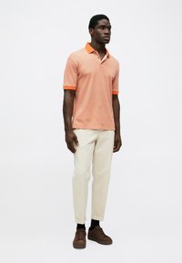 Cămașă polo cu mânecă scurtă, de culoare portocalie, cu un model geometric, având guler și manșete portocalii, asortată cu pantaloni crem și adidași maronii.